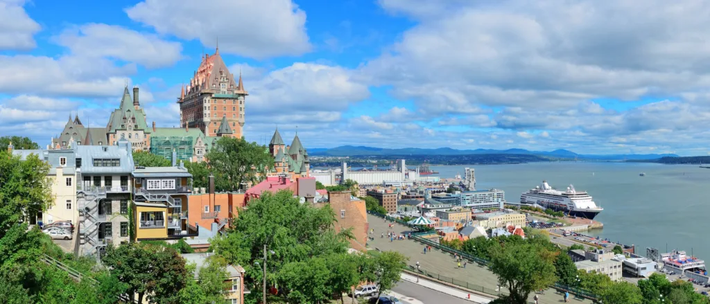 The Château Frontenac with its green copper roofs rising above the old town of Québec City, with the wide Saint Lawrence River and a cruise ship in port on the right, under a blue sky with clouds