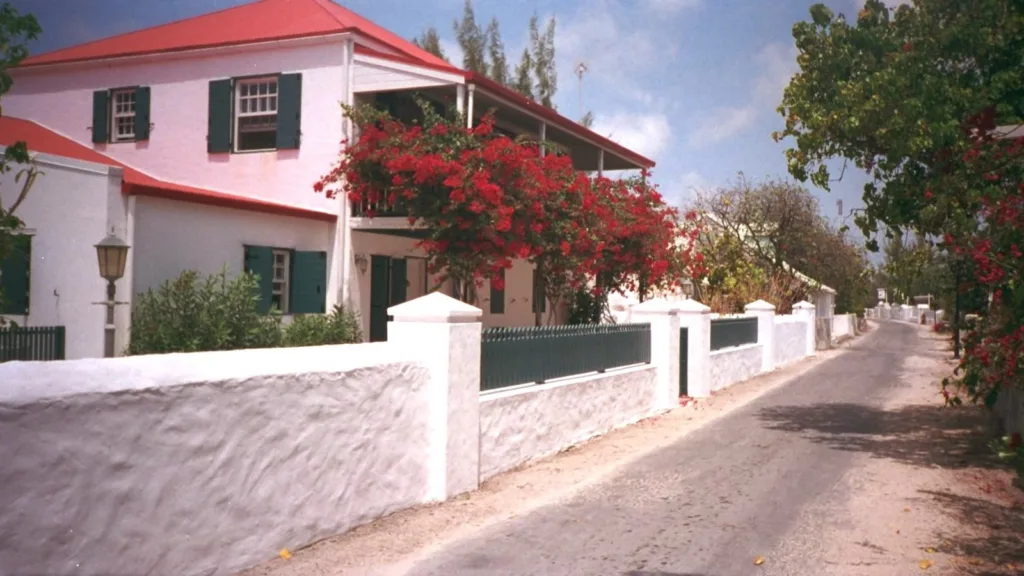 Whitewashed wall and fence beside a quiet street in Cockburn Town, Grand Turk, with a two-storey pink house under a red roof with green shutters and a red-flowering bougainvillea