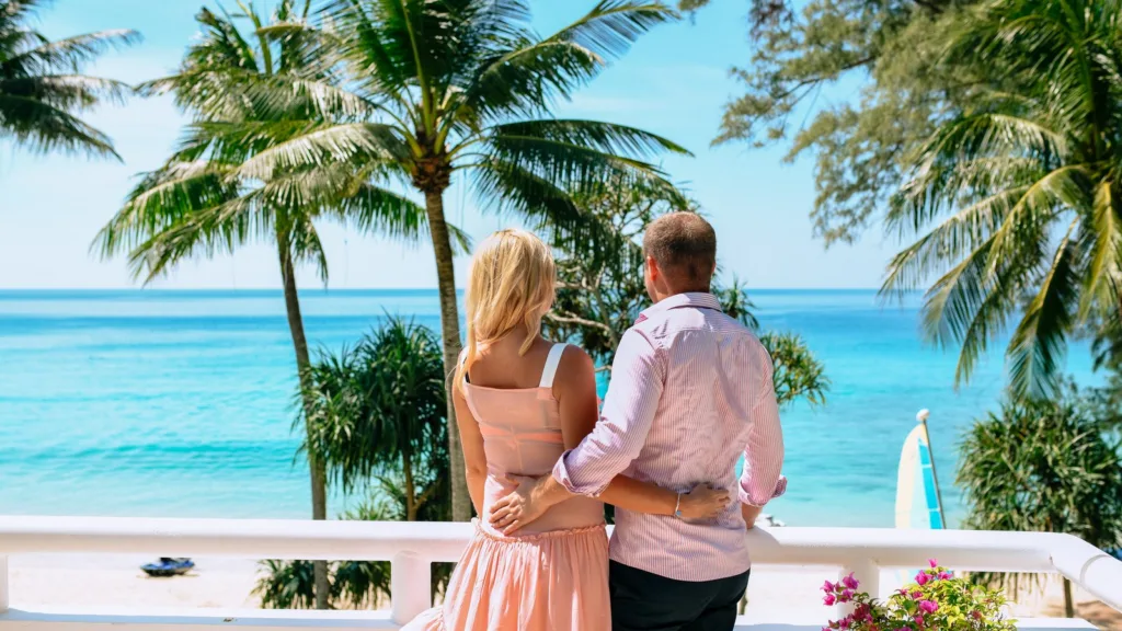 A couple seen from behind on a white balcony, looking out over palm trees to a turquoise sea and a sandy beach