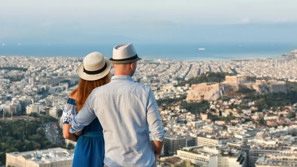 Một cặp đôi đội mũ cói nhìn từ đồi Lycabettus xuống các khối nhà ở Athens, về phía Acropolis và biển