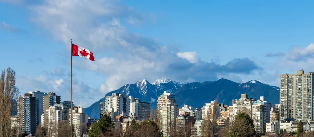 The Canadian flag flying on a pole in front of Vancouver high-rises, with snow-capped mountains behind under a blue sky