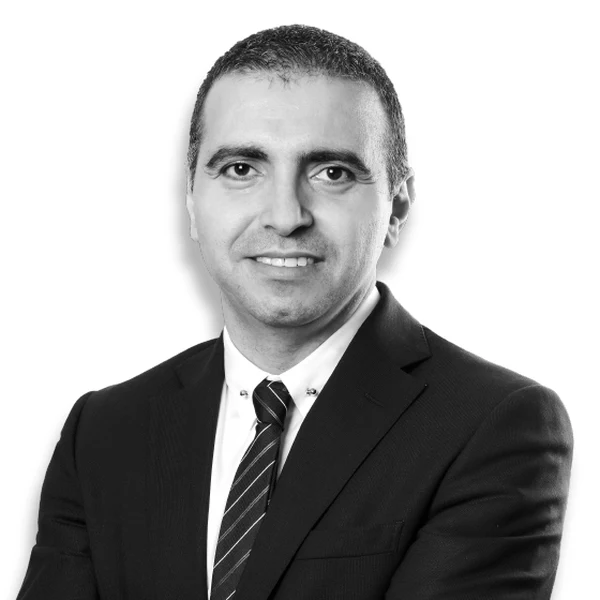 Black-and-white portrait of Evren Özmen in a dark suit, white shirt and striped tie, arms folded and smiling against a white background