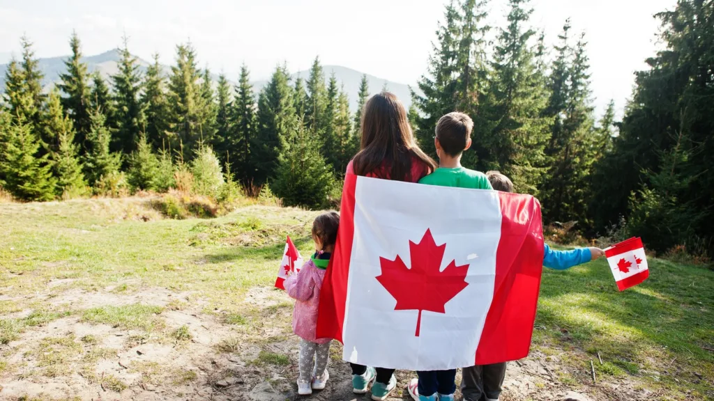 A mother and three children seen from behind, wrapped in a large Canadian flag and holding small flags, facing a forest and mountains