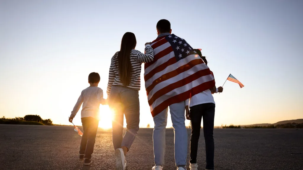 A family of four walking towards the sunset seen from behind, the father wrapped in a US flag and the children holding small flags