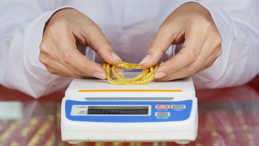 Two hands lay a gold chain on a white digital scale on a glass counter, with trays of gold jewellery blurred below