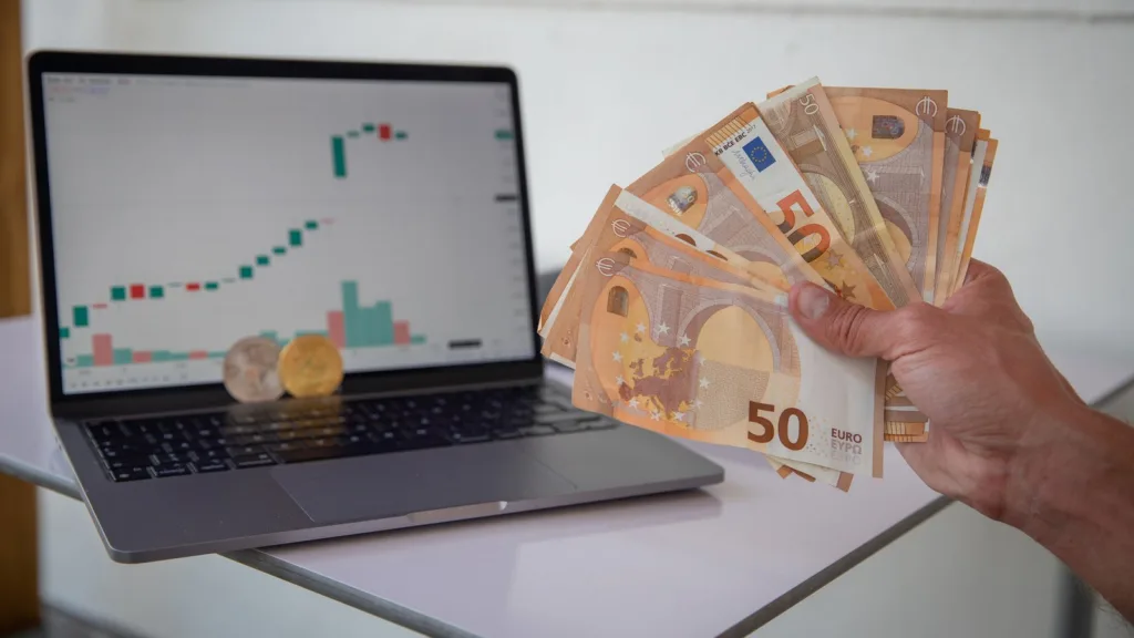 A hand holds a fan of €50 banknotes beside a laptop showing a candlestick chart, with two crypto coins standing in front of the screen