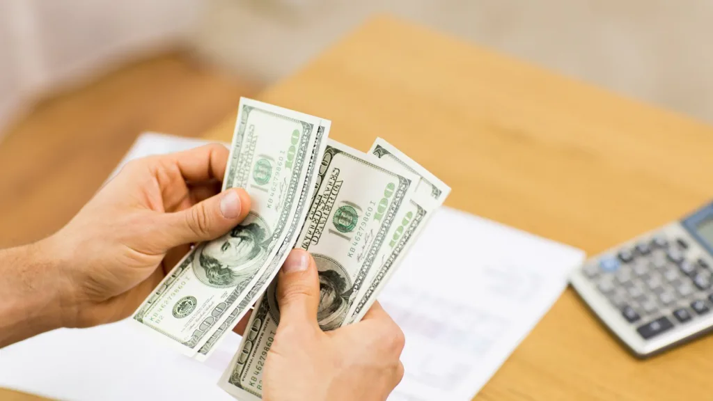 A man's hands count US$100 notes over a printed sheet on a wooden desk, with a calculator to the right
