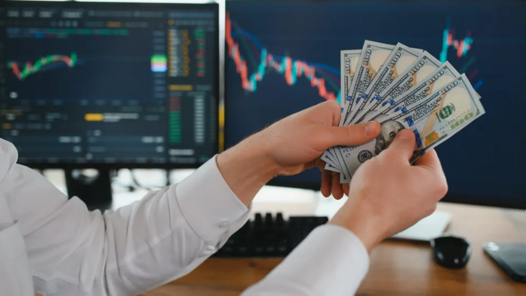 Hai bàn tay áo sơ mi trắng xòe một xấp tờ 100 đô-la Mỹ trước hai màn hình mờ hiển thị biểu đồ giá trên bàn gỗ