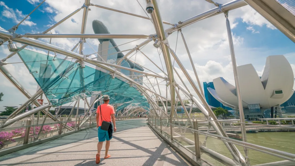 A man in a hat walks across the Helix Bridge in Singapore, with the ArtScience Museum and Marina Bay Sands behind