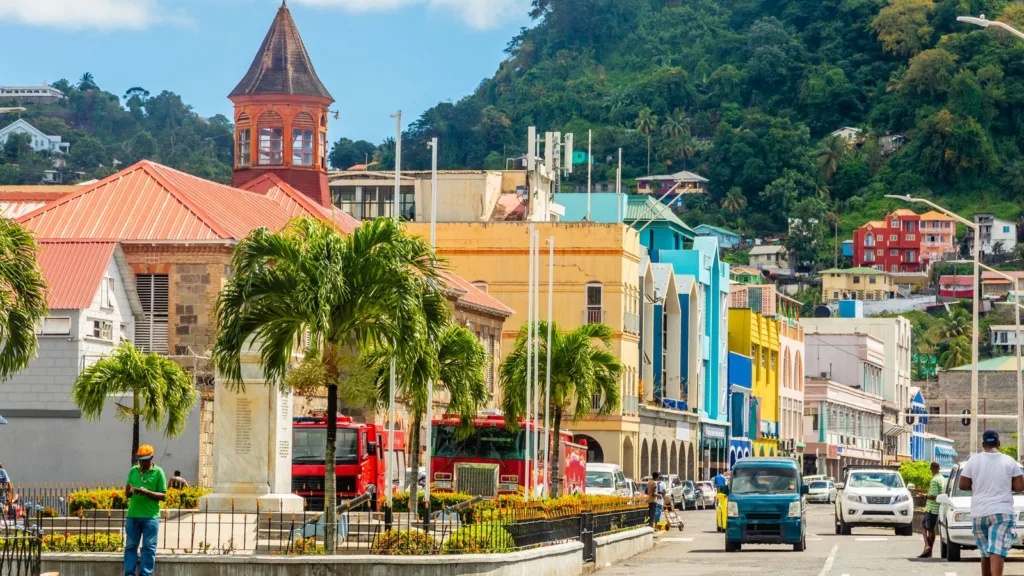 A street in central Kingstown, Saint Vincent, with a red-roofed stone building and small bell tower, two red fire engines parked behind a white memorial, palm trees, pedestrians and brightly painted buildings below a green hillside