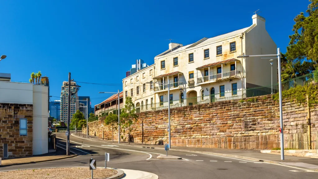 Three-storey sandstone terraces with iron balconies above a stone retaining wall in Millers Point, Sydney, with city towers behind
