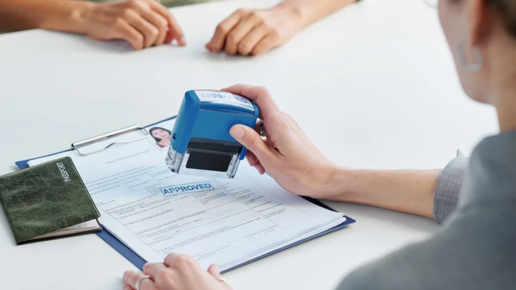 An official in glasses holds a blue stamp over a visa application form beside a closed green passport, across a white desk from an applicant whose hands rest on the table