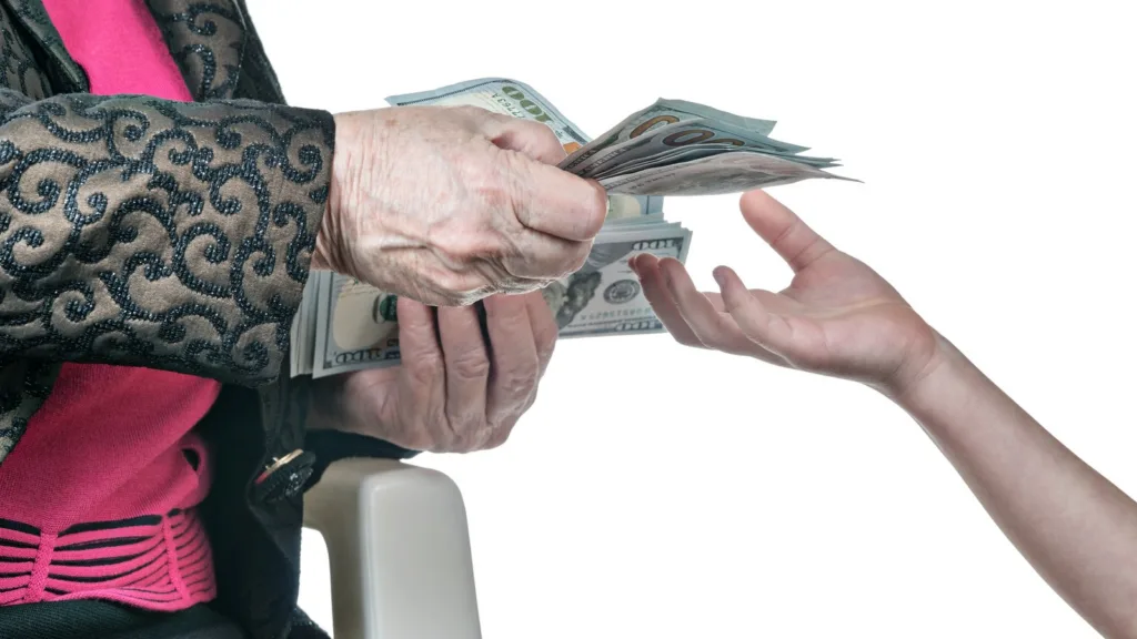 An older woman's wrinkled hands hold out a fan of US$100 notes towards a younger person's open hand, against a white background