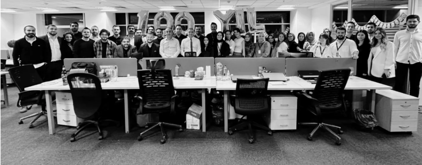 Black-and-white photo of about forty staff standing behind rows of desks in an open-plan office, with 100 YIL balloons behind them