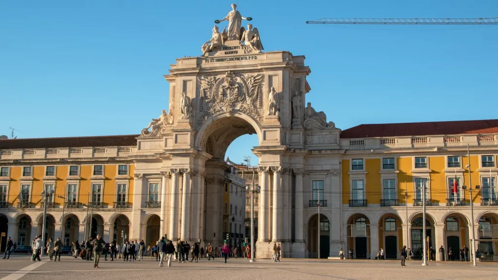 People cross Praça do Comércio in Lisbon in low sunlight, beneath the Rua Augusta Arch and the square's yellow arcaded buildings