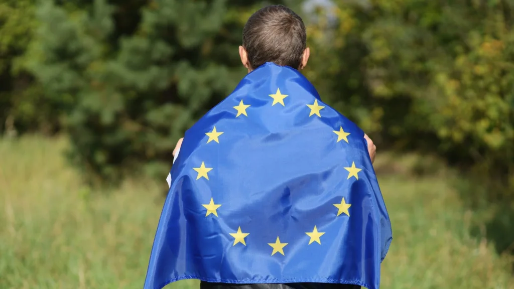 A person seen from behind with the European Union flag draped over the shoulders, standing in a grassy field by trees