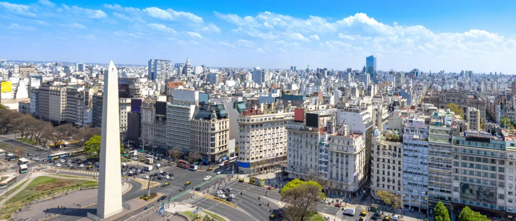 Đài tưởng niệm Obelisco giữa đại lộ 9 de Julio ở Buenos Aires nhìn từ trên cao, hai bên là các dãy nhà trung tâm thành phố dưới trời xanh nhiều mây trắng