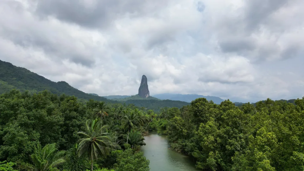 Đỉnh núi đá Pico Cão Grande nhô lên giữa rừng nhiệt đới ở São Tomé, phía trước là dòng sông và hàng cọ dưới trời nhiều mây