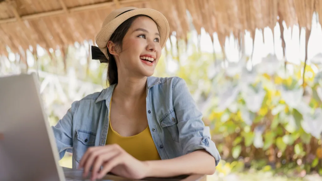 A smiling woman in a straw hat and denim shirt types on a laptop under a thatched roof, with tropical plants behind her