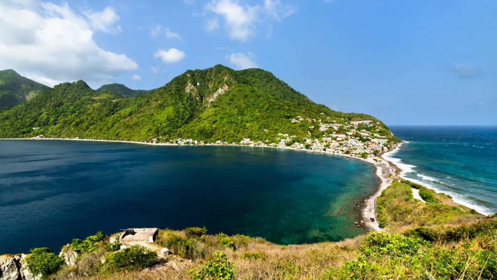 View over the bay and the isthmus at Scotts Head, Dominica, with a green wooded mountain, coloured houses along a pebble shore and dark blue sea under a blue sky