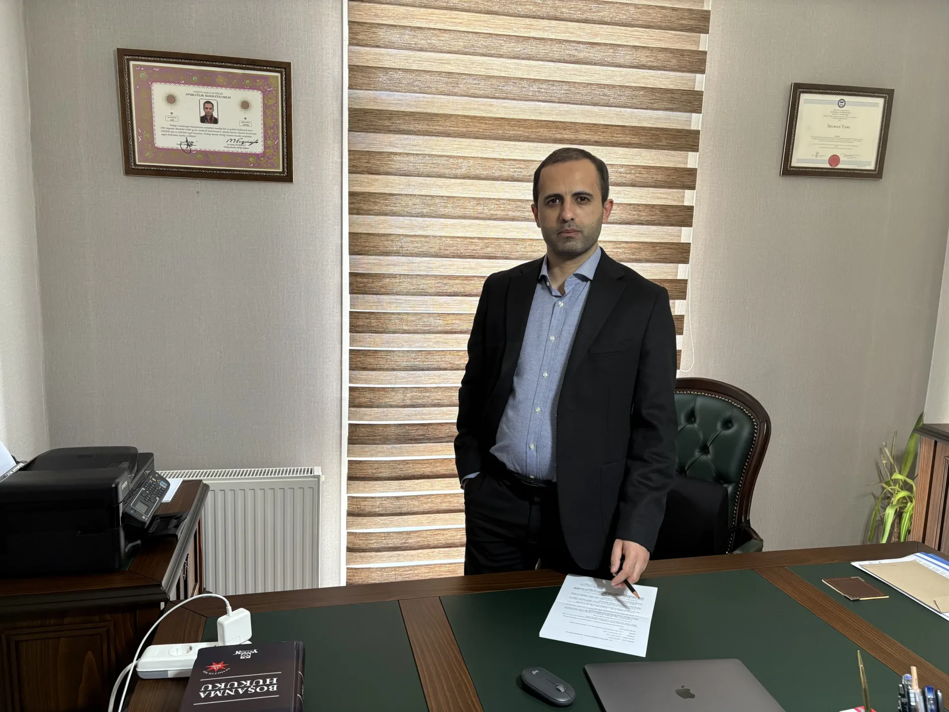 Lawyer Selman Yanı in a black jacket and blue shirt standing behind his desk, with framed certificates on the walls either side