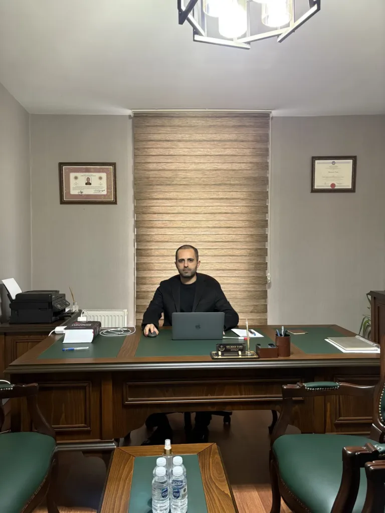 Selman Yanı seated at a wooden desk with a green leather top, framed certificates on the walls either side, green leather chairs and a coffee table in front