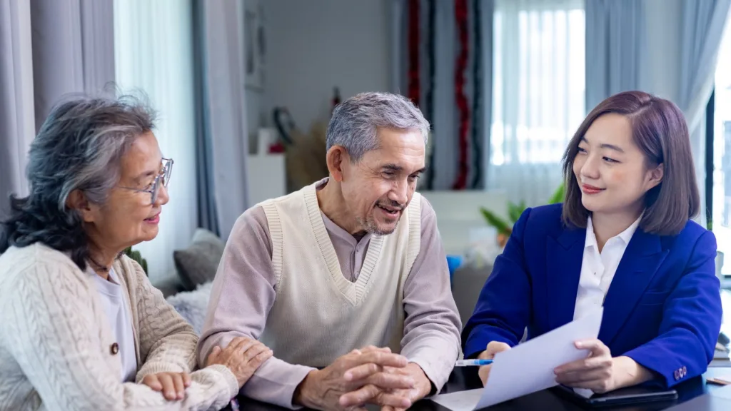 An older Asian couple at a dining table listen as a woman in a blue blazer goes through documents with them