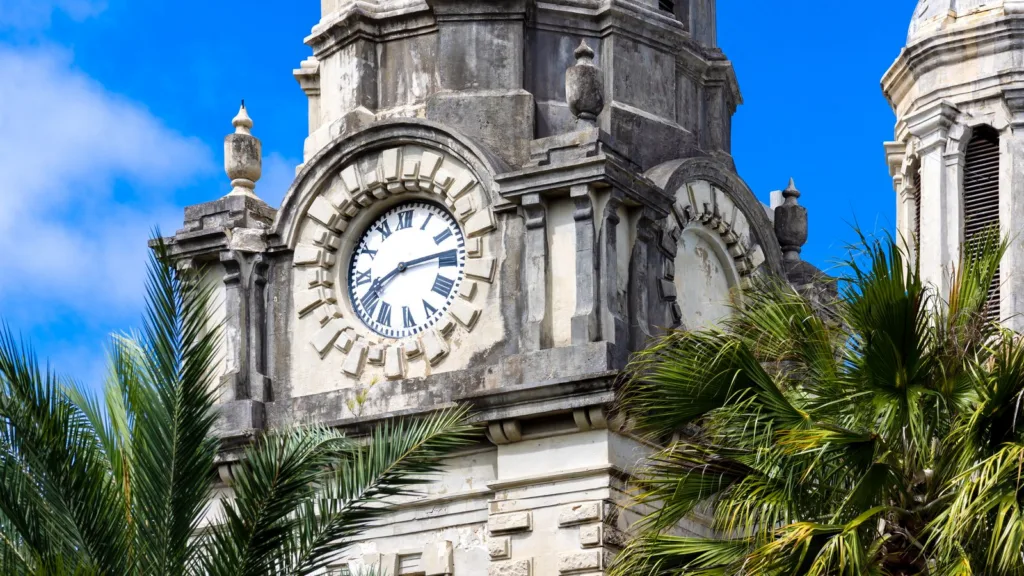 The clock tower of St John's Cathedral in St John's, Antigua, with a white clock face and Roman numerals set in weathered pale stone, between palm fronds under a blue sky