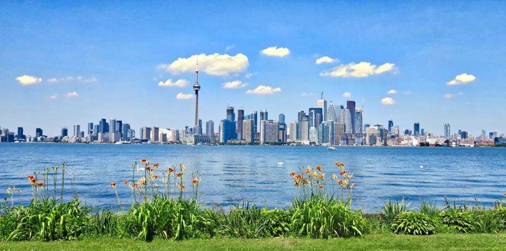 The Toronto skyline and CN Tower seen across Lake Ontario from the Toronto Islands, under a blue sky with white clouds and orange lilies along the shore