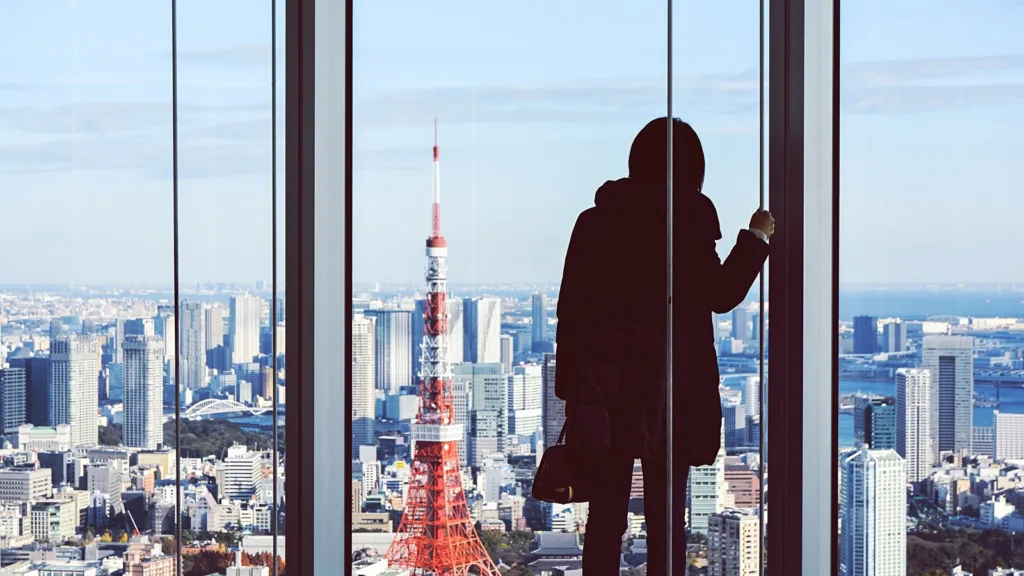 A person in silhouette stands at a floor-to-ceiling window high above Tokyo, with the red and white Tokyo Tower and the bay in view