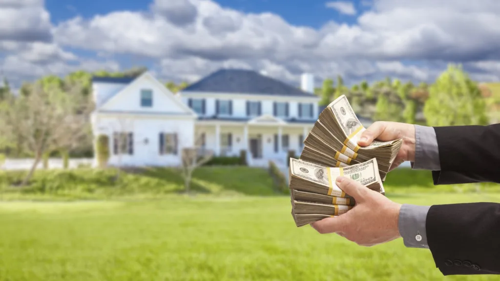 Two hands fan out bundles of US$100 notes in front of a white two-storey house with a porch and a wide lawn