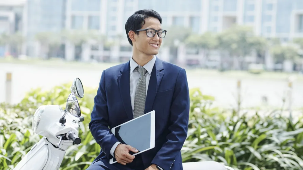 Một thanh niên mặc vest xanh đậm, đeo kính, ngồi trên chiếc xe tay ga màu trắng, tay cầm máy tính bảng, phía sau là dòng sông và các tòa nhà văn phòng