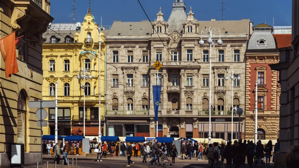 Pedestrians crossing Ban Jelačić Square in Zagreb in spring sunshine, with market stalls under blue awnings in front of ornate yellow and stone facades